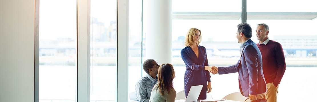 A group of 5 professionals sit in a glass-walled conference room, with two shaking hands.