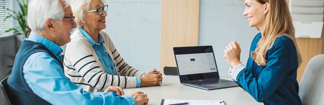 A couple sits across from their accountant at a desk reviewing documents and a laptop screen during a tax‑planning meeting.